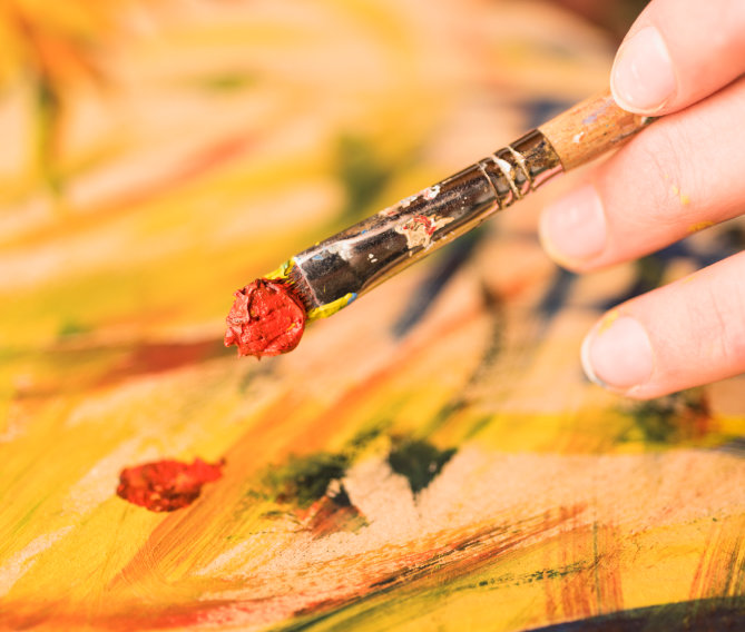 Hand hält Pinsel mit roter Acrylfarbe auf gelber Leinwand